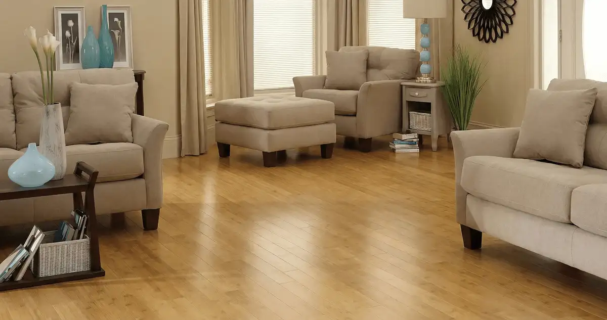 A living room with professionally installed bamboo flooring, beige sofas, a matching ottoman, a side table with a blue lamp, vases, framed pictures, and large windows with blinds and curtains in Cincinnati, OH